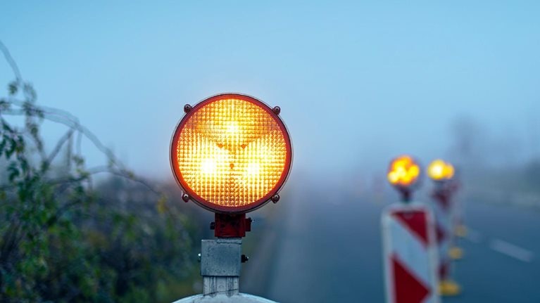 A bright amber warning light sits atop a road barrier, glowing vividly against a foggy, muted background. Additional blurred warning lights and striped barriers line the roadside.