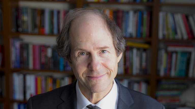 Portrait of Cass Sunstein in front densely filled bookcases.