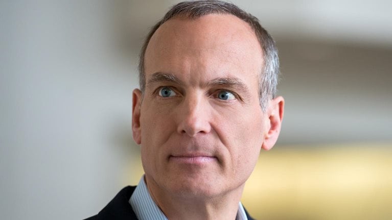 Glenn Fogel with short salt and pepper hair and blue eyes, wearing a dark blazer over a light collared shirt. He is looking slightly off to the side with a neutral, thoughtful expression against a softly blurred indoor background.