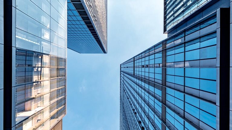 A dramatic upward view between modern glass skyscrapers, their reflective blue surfaces forming a geometric frame around the sky.