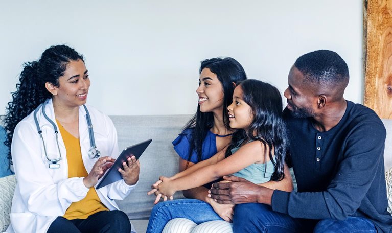 A hispanic doctor sitting on a couch talking to a mixed-race family during a home visit.