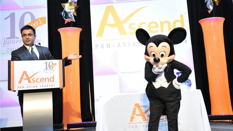 Vivek Sharma stands at a podium onstage and introduces a person in a Mickey Mouse costume