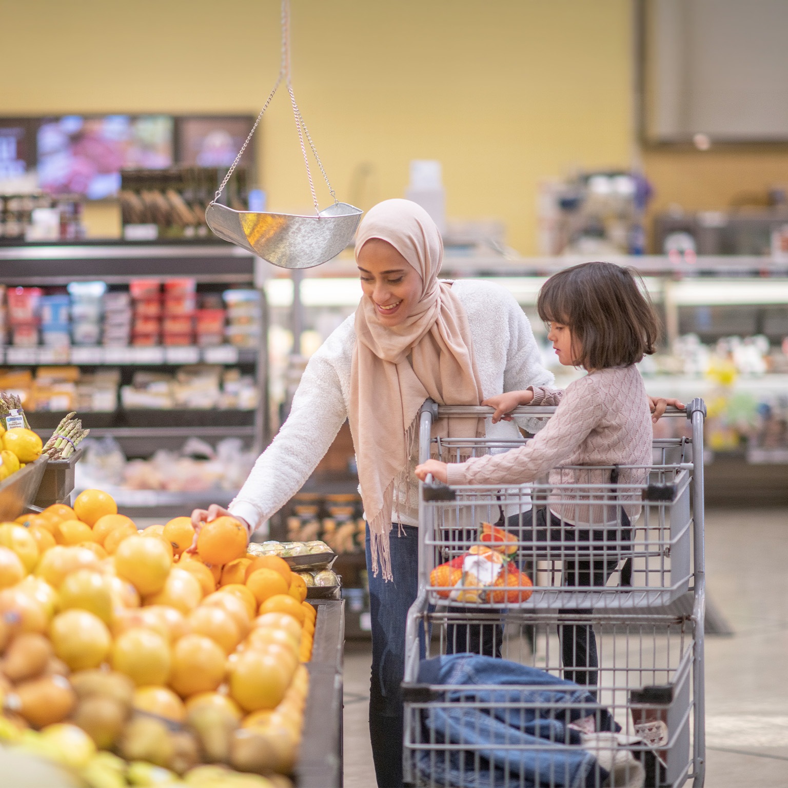 A Muslim mother goes grocery shopping with her daughter. Her daughter is sitting on the grocery cart while she picks through some fresh produce.
