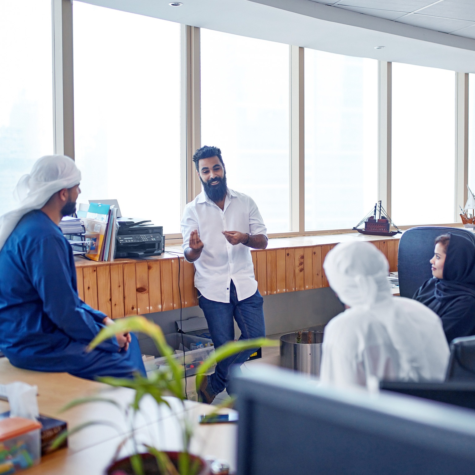 A young businessman in jeans and a button down sharing ideas with male and female colleagues in traditional Emirati clothing. The team is casually seated in a circle in a open office setting.