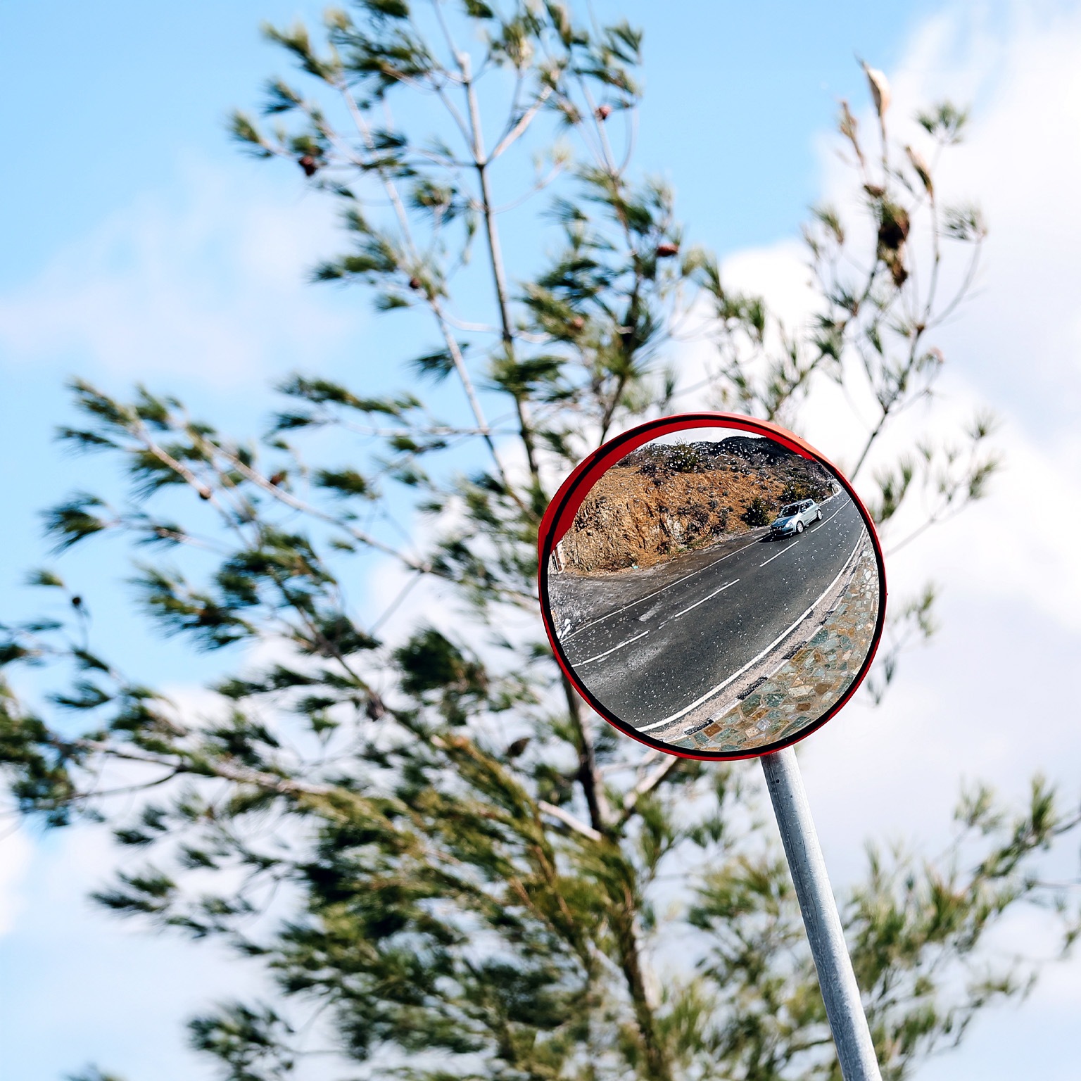 A round roadside convex mirror mounted on a pole, reflecting a winding road with a car driving along it. Behind the mirror, tree branches sway against a bright blue sky with scattered clouds.