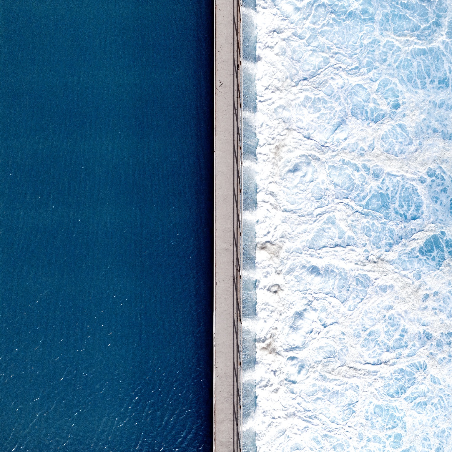 An overhead shot captures the stark contrast between deep, tranquil blue water on one side and churning, white-foamed turquoise waves on the other, bisected by a narrow concrete pier. The smooth, dark surface on the left stands in dramatic opposition to the textured, frothy turbulence crashing against the right side of the man-made barrier.