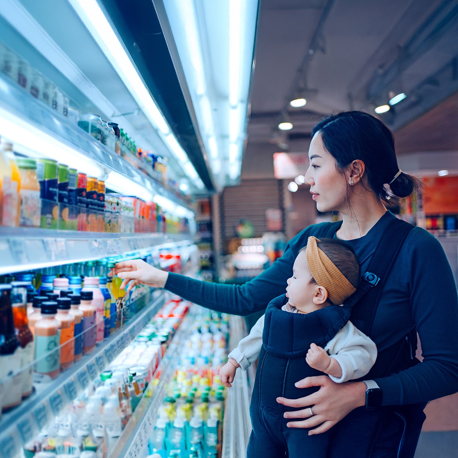 Young Asian mother carrying her baby girl in a baby carrier, doing grocery shopping in supermarket. Choosing fresh organic fruit juice for her family. Healthy eating habits with balanced diet.