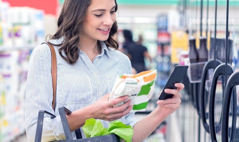 Confident young Hispanic woman holds smart phone as she reads a nutrition label on a bag of frozen vegetables. She is holding a shopping basket filled with healthy foods.
