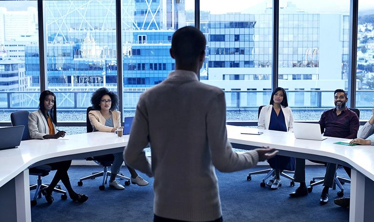 Image of a man standing at the head of a curved white table in a modern office, addressing diverse professionals in a meeting.