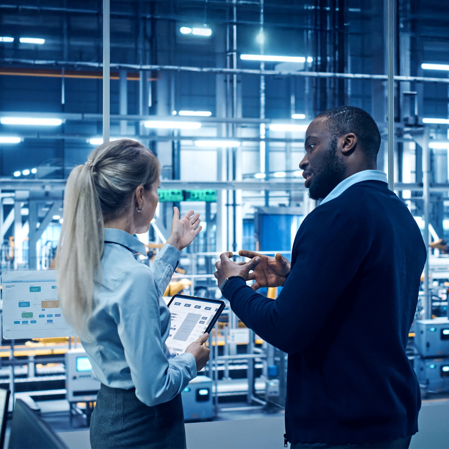 Two engineers stand in a high-tech automotive manufacturing facility discussing data displayed on digital screens and a tablet, with robotic machinery operating in the background.