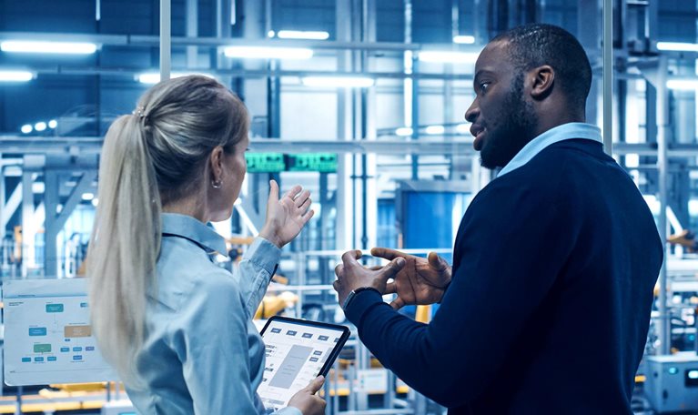 Two engineers stand in a high-tech automotive manufacturing facility discussing data displayed on digital screens and a tablet, with robotic machinery operating in the background.