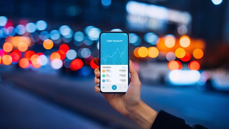 Woman's hand holding up smartphone against illuminated city street lights, using NFT investment wallet on smartphone in city, working with blockchain technologies.