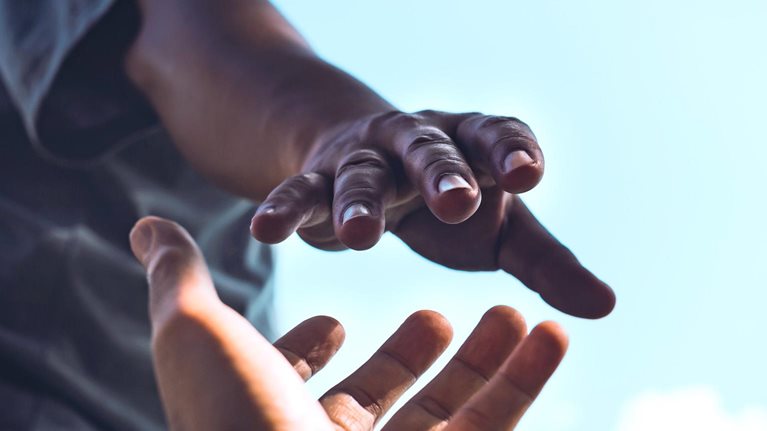 A tightly cropped image of a person reaching down with an outstretched hand while another person reaches upward, their fingers almost touching. Set against a bright sky, it conveys a strong sense of help, connection, and support.
