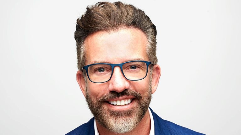 Nick Jeffery smiling warmly at the camera, wearing glasses and a blue suit jacket over a white shirt. He has neatly styled brown hair and a trimmed salt-and-pepper beard, set against a clean, light background.
