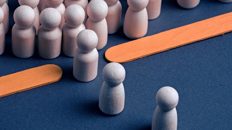 Wooden peg figures arranged on a dark blue surface, with a dense group in the background and several individual figures in the foreground. Two flat orange wooden sticks lie diagonally creating a bottleneck between the two groups.
