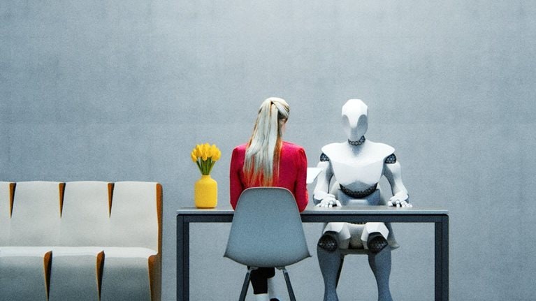 A woman in a red jacket sitting at a table across from a humanoid robot in a sparsely decorated office space.