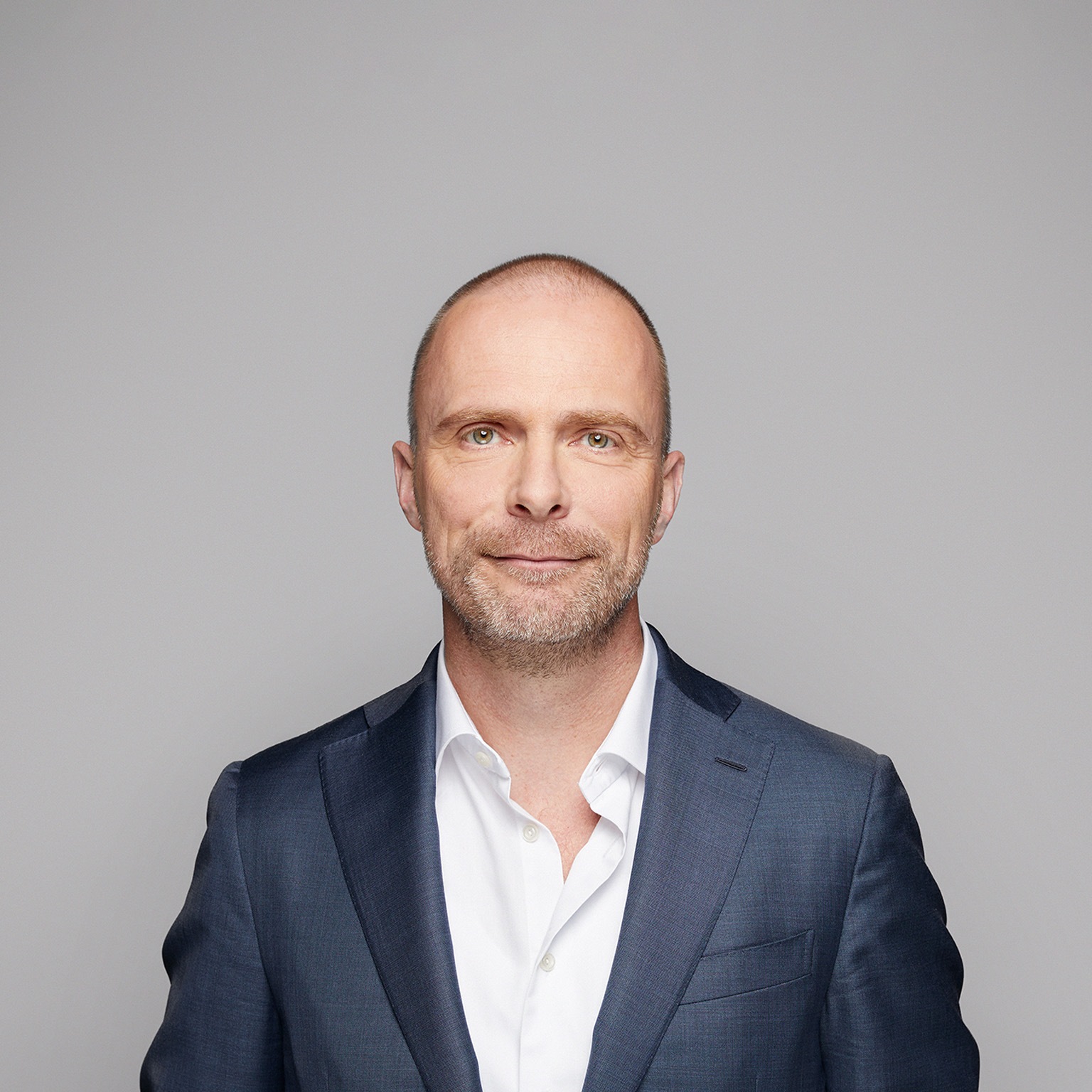 A portrait of Robert Carsouw, with a light beard, and a easy smile. He is wearing a dark blue suit and a white, open-collared shirt against a light gray background.