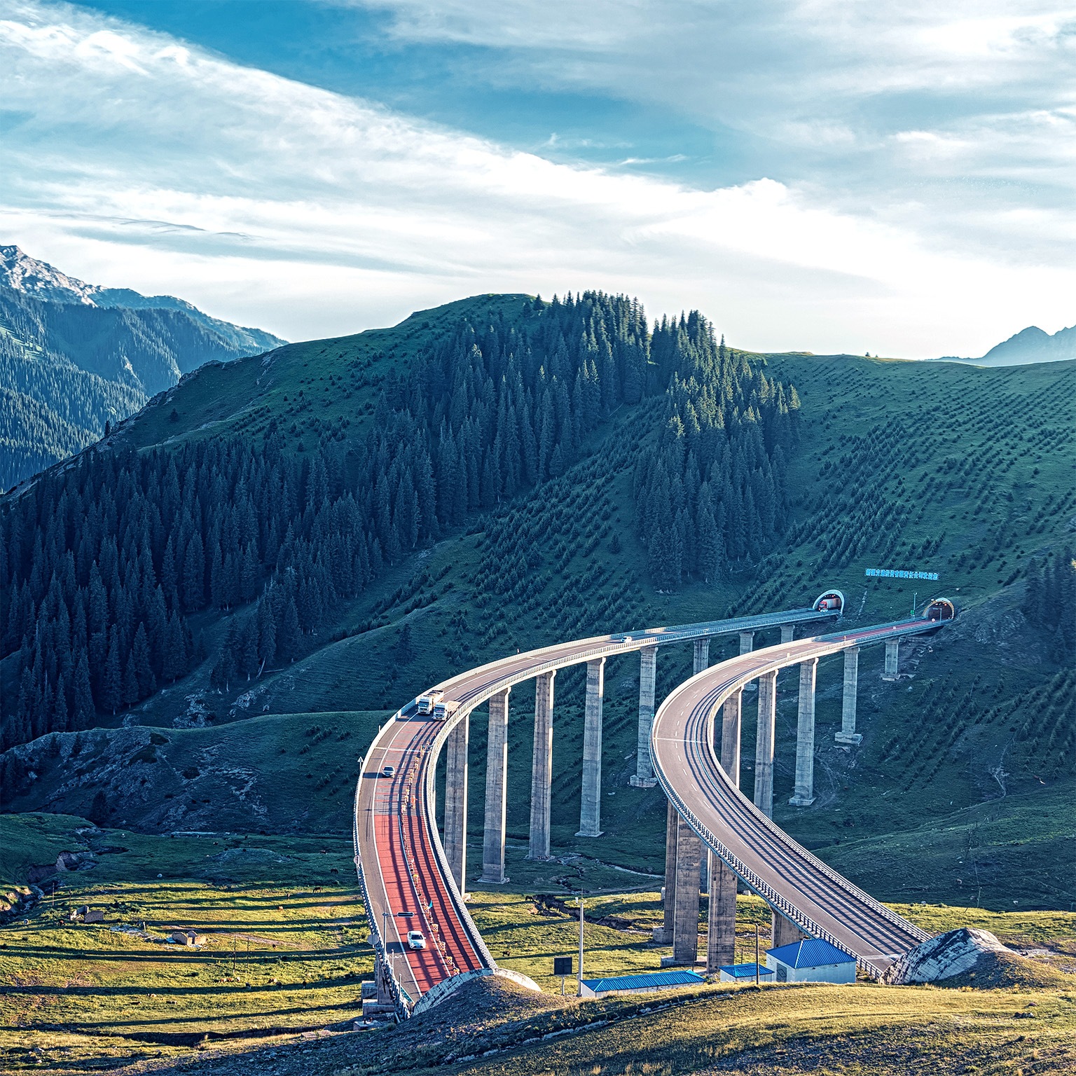 A winding elevated highway curves gracefully through a lush green mountainous landscape, supported by tall pillars. In the distance, forested hills and rugged peaks stretch beneath a bright, partly cloudy sky.