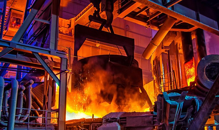 The interior of a large industrial steel plant, where molten metal glows bright orange amid heavy machinery and towering metal structures.