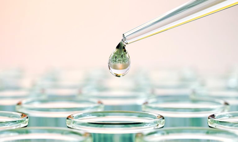 A close-up of a laboratory pipette releasing a droplet of liquid above a row of clear test tubes filled with fluid.