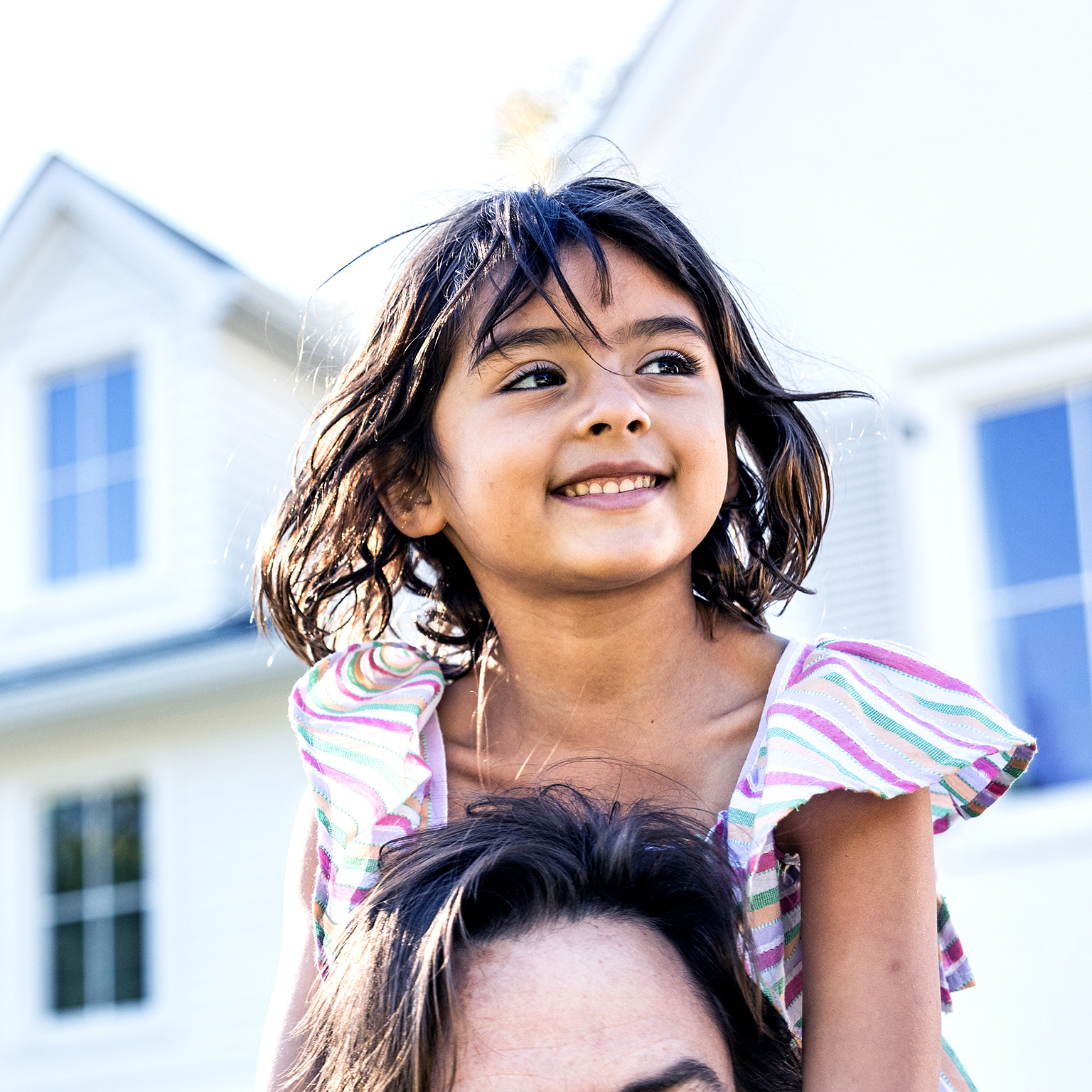 Young girl riding on her fathers shoulders in front of suburban home