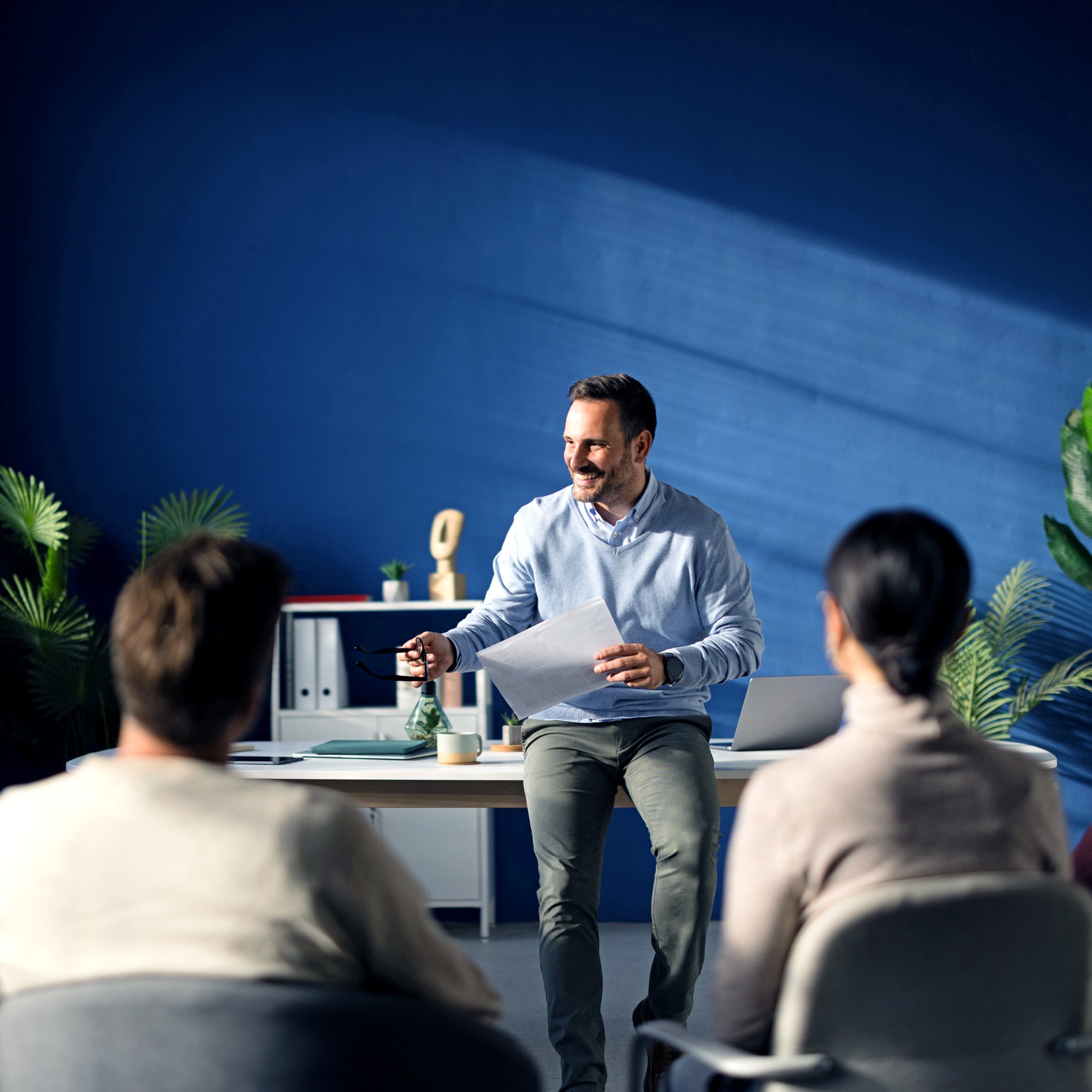 A cheerful CEO stands beside his desk, attentively hearing out a diverse group of employees seated in chairs.