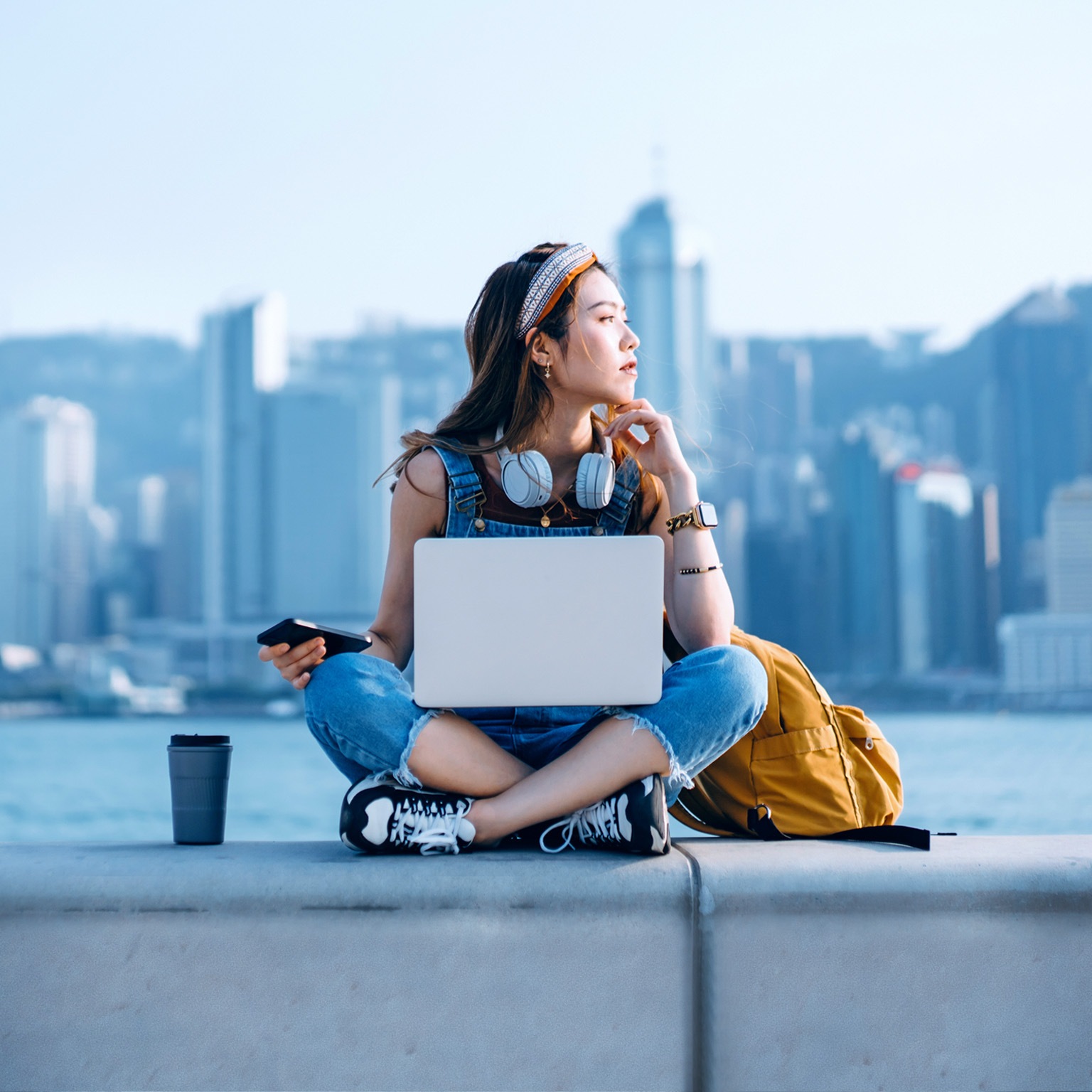 A young person, with a backpack, a beverage tumbler, and a laptop, is sitting on a ledge with back to the waterbody against the backdrop of skyscrapers.