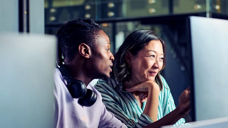 Two colleagues, a young Black man and an Asian woman, work side by side at a computer, intently looking at the screen.