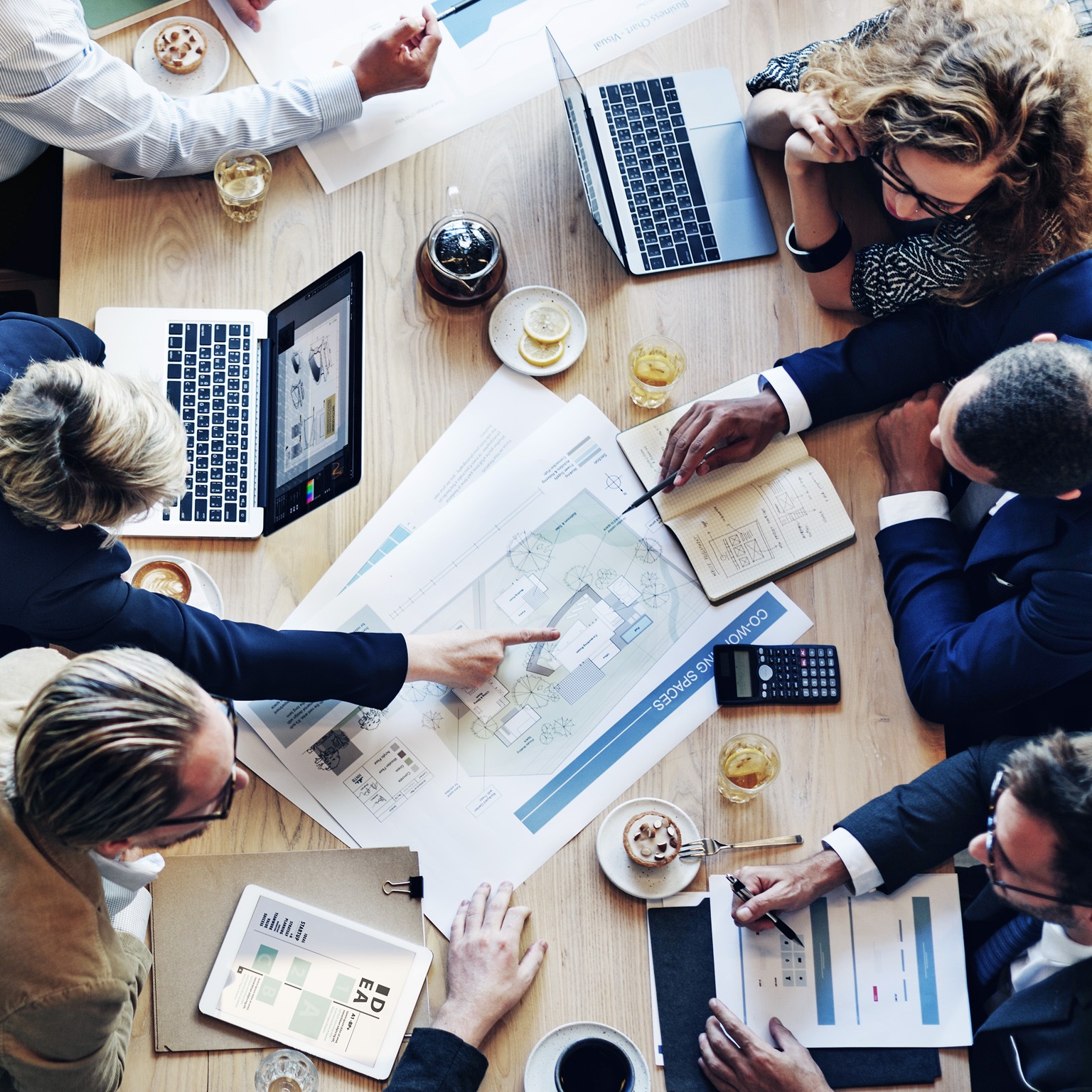 From a bird's-eye view, a diverse group of business professionals is gathered around a table covered with laptops, tablets, design plans, and financial reports The team is engaged in a spirited conversation about a project.