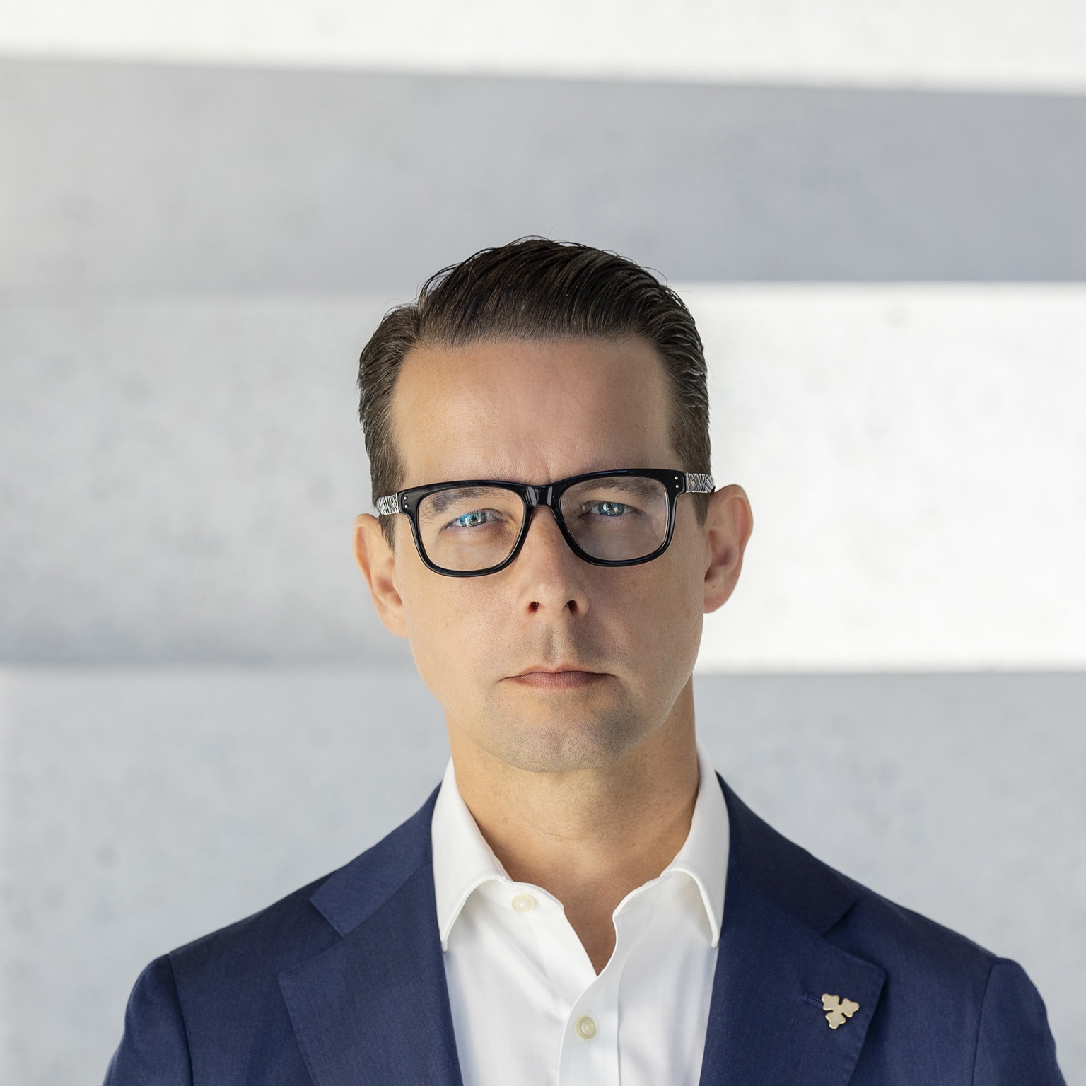 Jacob Aarup-Andersen is depicted in a portrait with a stern look, wearing glasses, a deep blue suit coat, and a white button-down shirt.