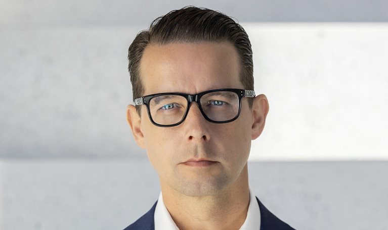 Jacob Aarup-Andersen is depicted in a portrait with a stern look, wearing glasses, a deep blue suit coat, and a white button-down shirt.