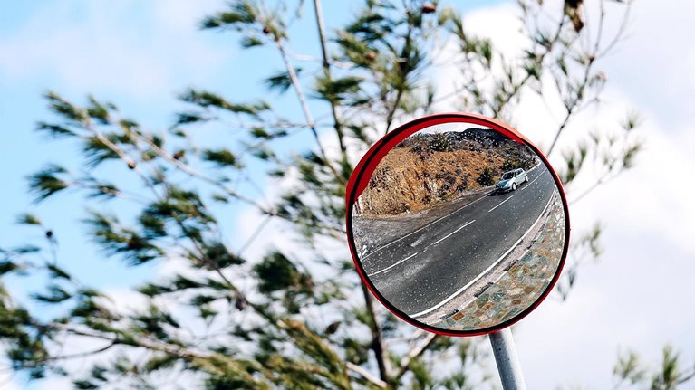 A round roadside convex mirror mounted on a pole, reflecting a winding road with a car driving along it. Behind the mirror, tree branches sway against a bright blue sky with scattered clouds.