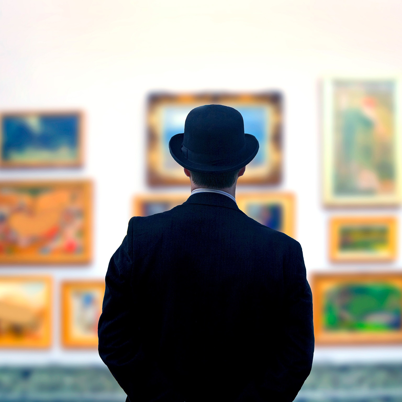 A man in a black suit and bowler hat looks at art in a gallery.