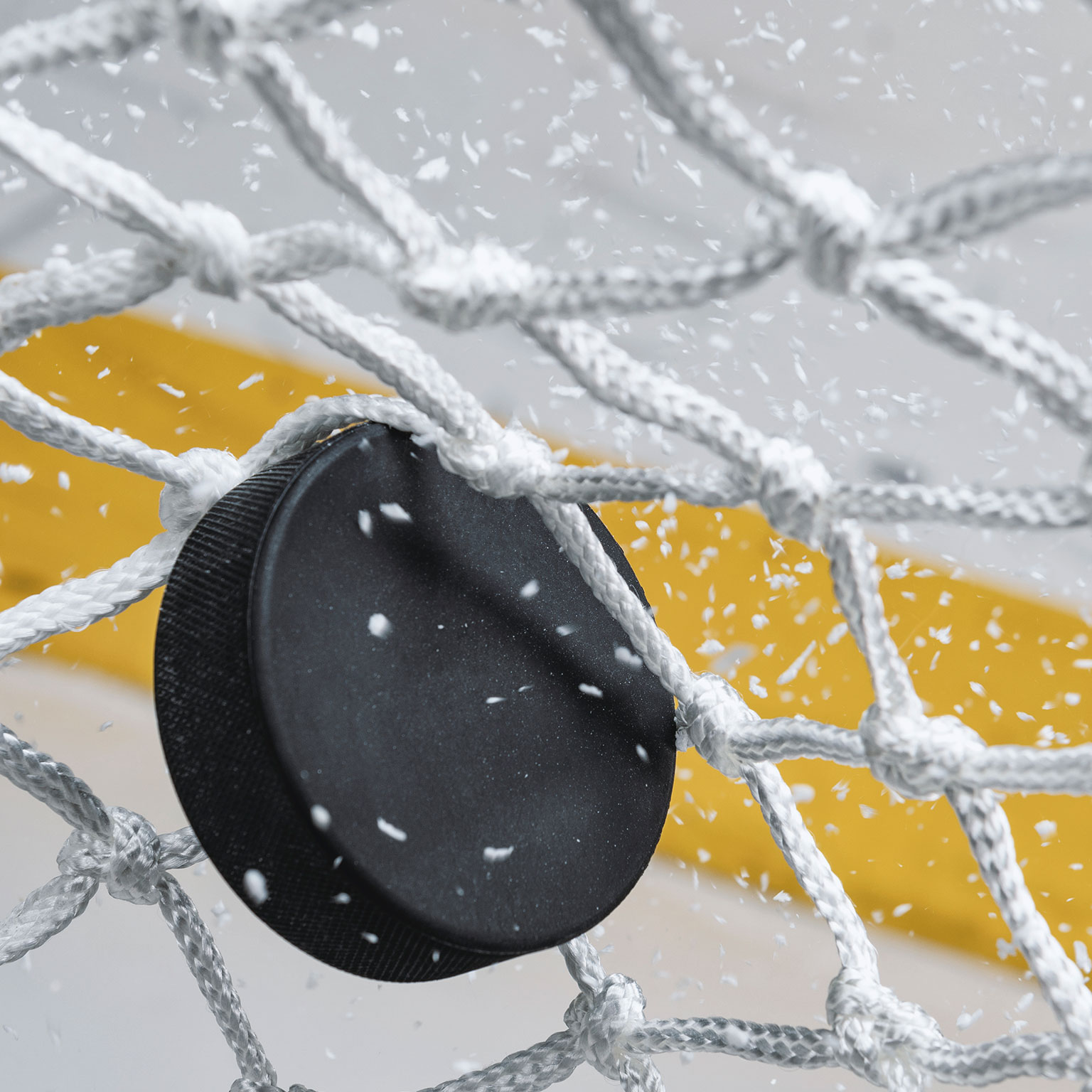 A close-up view of an Ice Hockey puck hitting the back of the goal net as shavings fly by, viewed from the front. Scoring a goal in ice hockey.