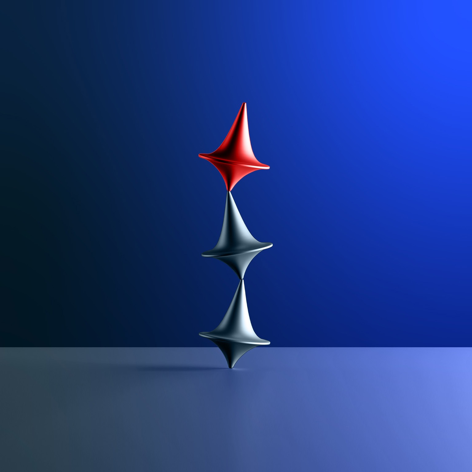 Three tops, two silver and one red, are arranged in a vertical stack on a blue surface. The tops are uneasily balanced, with the red top positioned above the two silver ones.