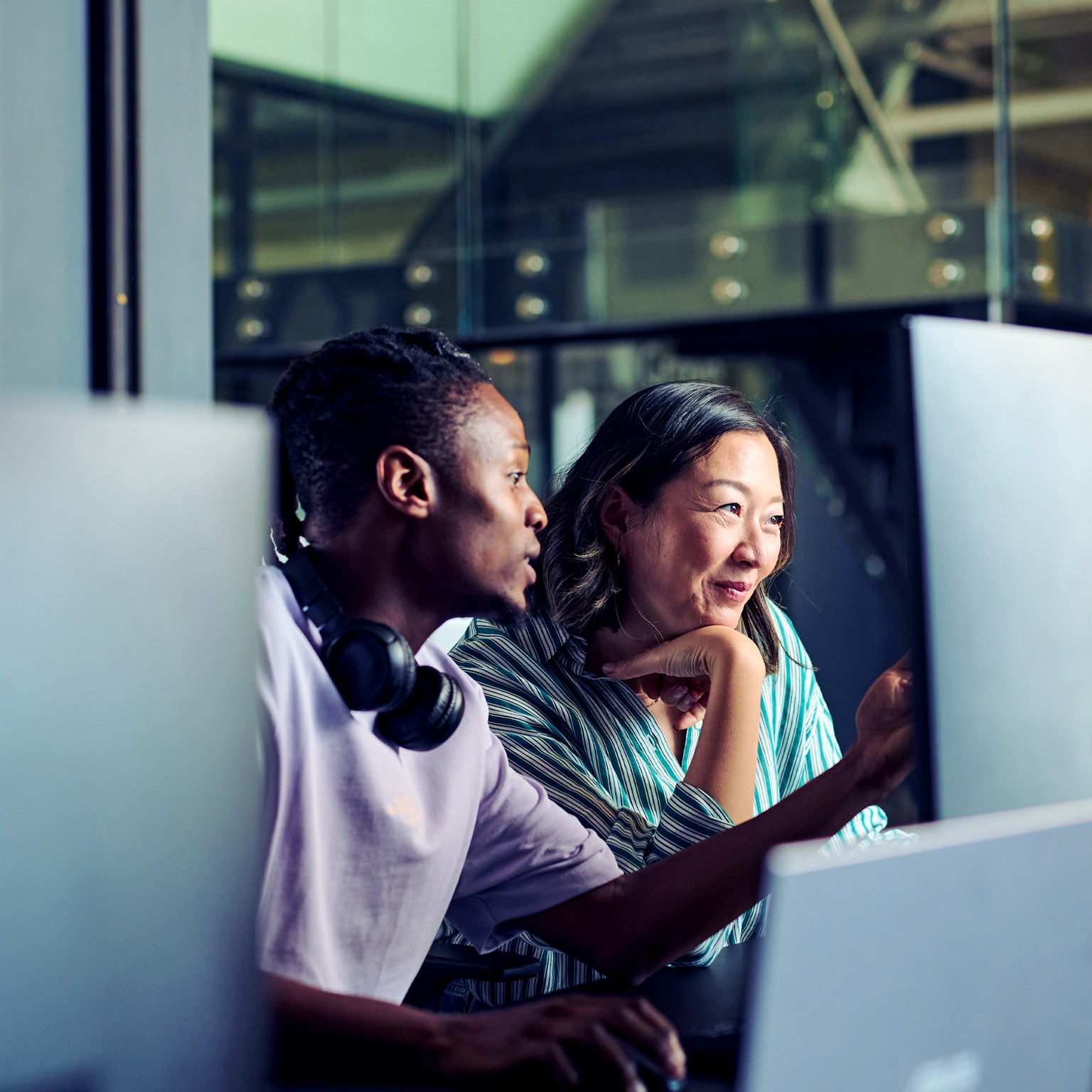 Two colleagues, a young Black man and an Asian woman, work side by side at a computer, intently looking at the screen.