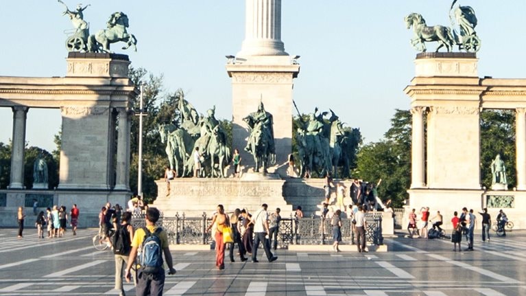 Visitors strolling and capturing images at Heroes Square in Budapest, with the Millennium column prominently displayed at the center of the view.