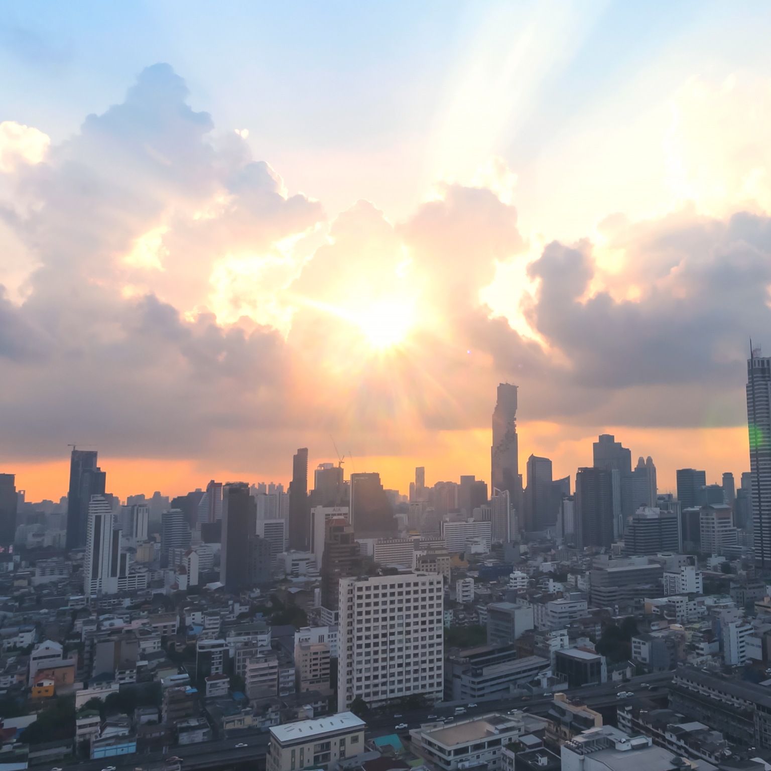 The image is an aerial view of the Bangkok skyline, likely near the Sathorn district, featuring several modern skyscrapers at sunset or sunrise. The sun is low on the horizon, casting a warm orange glow and dramatic light rays through the clouds.