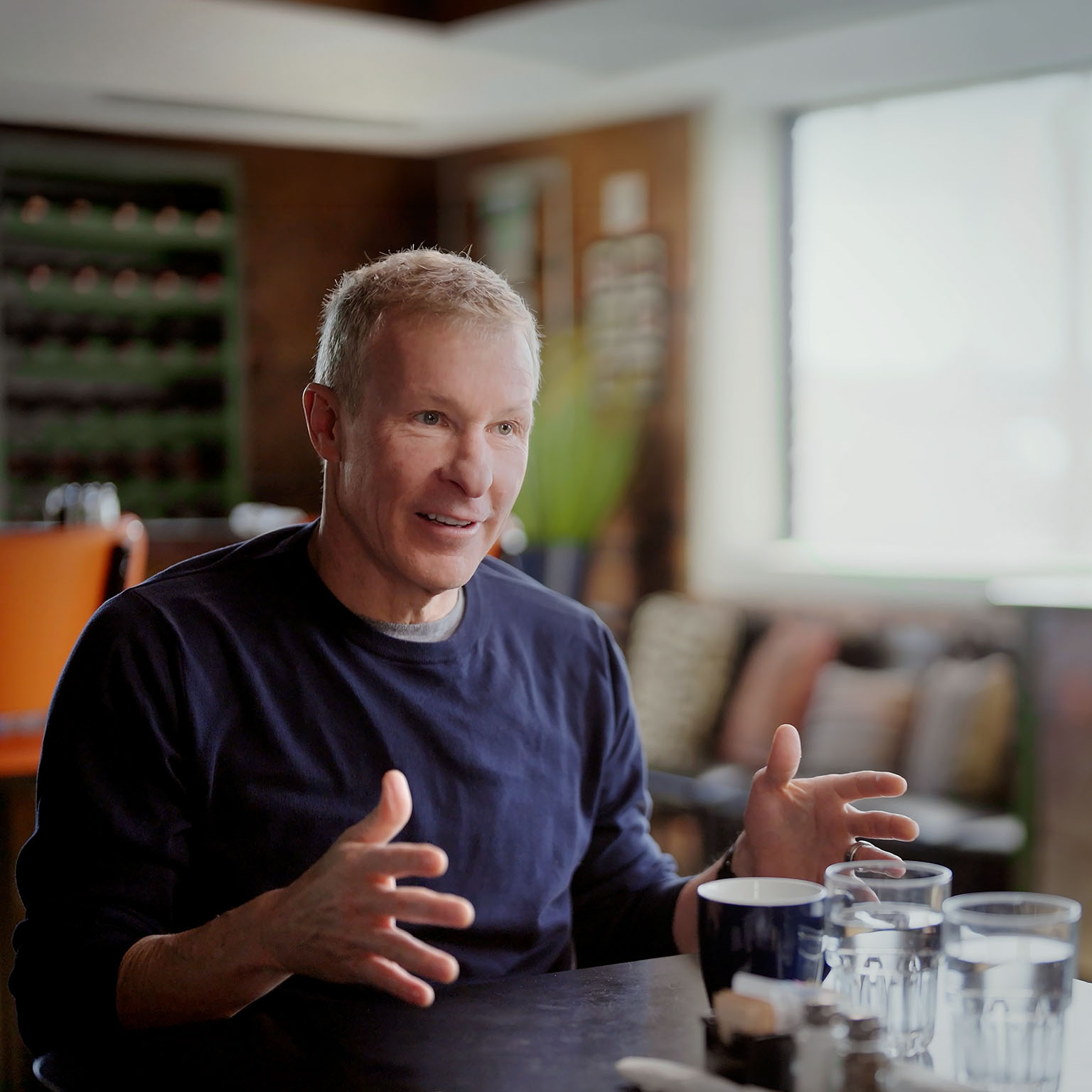 A middle-aged man gesturing while speaking at a table with mugs and glasses in a cozy, well-lit room.