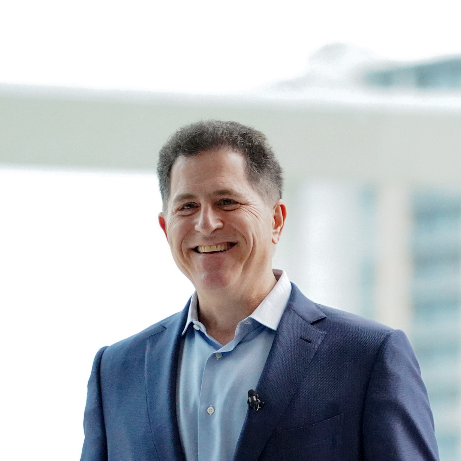Michael Dell, wearing a blue suit jacket and smiling in an office filled with natural sunlight.