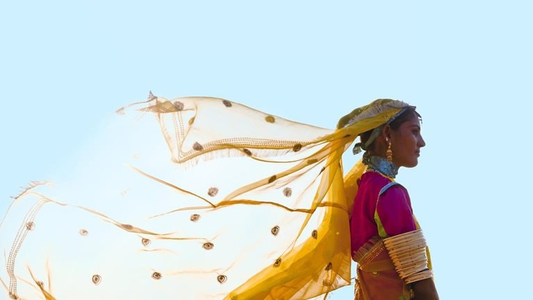 A woman from India stands tall and proud in a vibrant saree, her yellow veil fluttering in the wind against a backdrop of a clear blue sky.