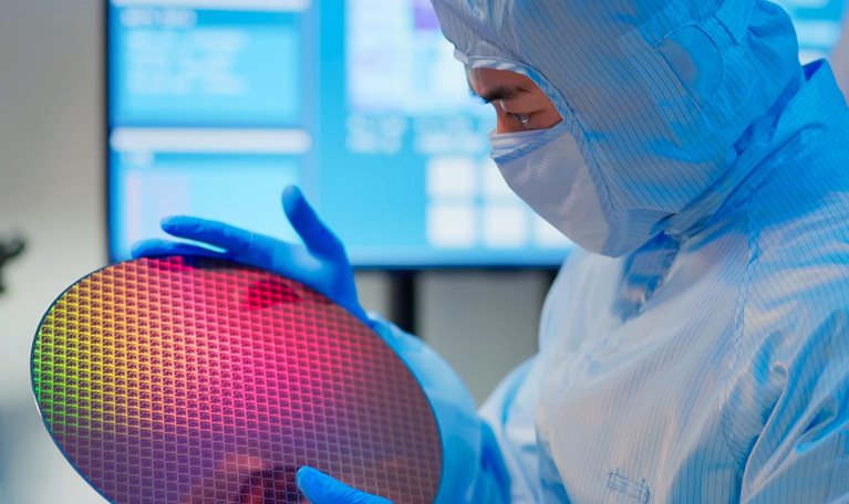 Male technician in sterile coverall holds wafer that reflects many different colors with gloves and check it at semiconductor manufacturing plant.