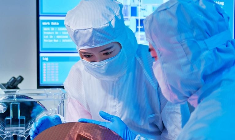 Two Asian technicians in sterile coverall hold wafer with gloves and check it at semiconductor manufacturing plant