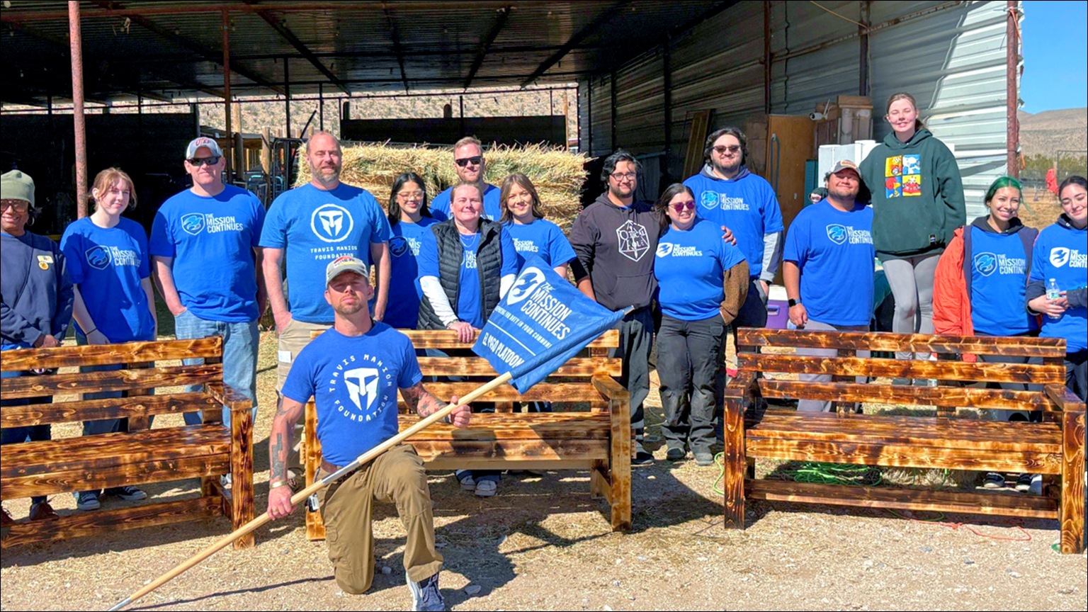 The Mission Continues’s volunteers posing outdoors behind newly built wooden benches, with one person kneeling in front holding a flag.