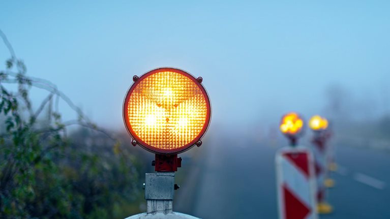 A bright amber warning light sits atop a road barrier, glowing vividly against a foggy, muted background. Additional blurred warning lights and striped barriers line the roadside.