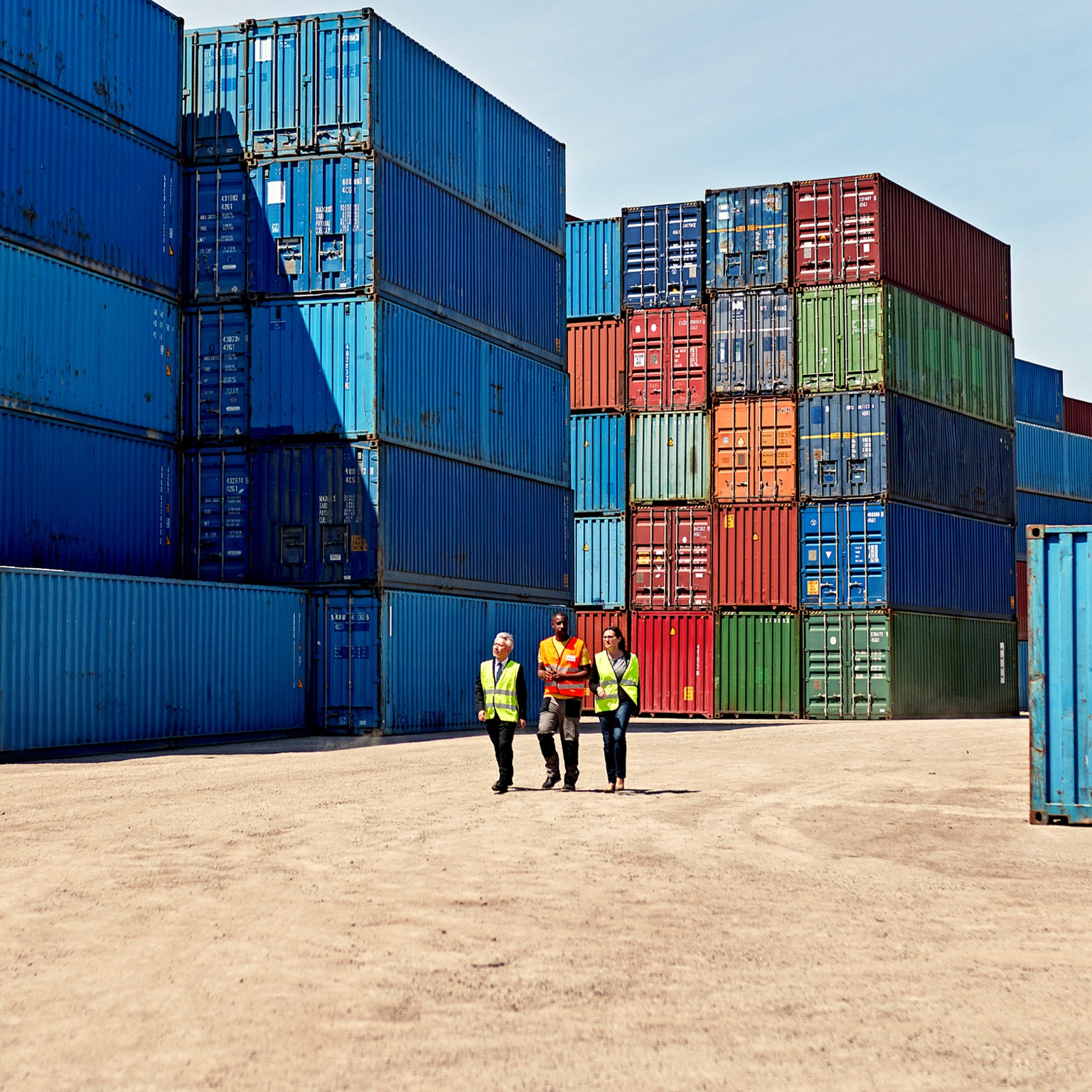 Three people in high-visibility vests walking in a shipping yard, amidst stacks of colorful shipping containers. The scene suggests a meeting in a bustling port or logistics area.