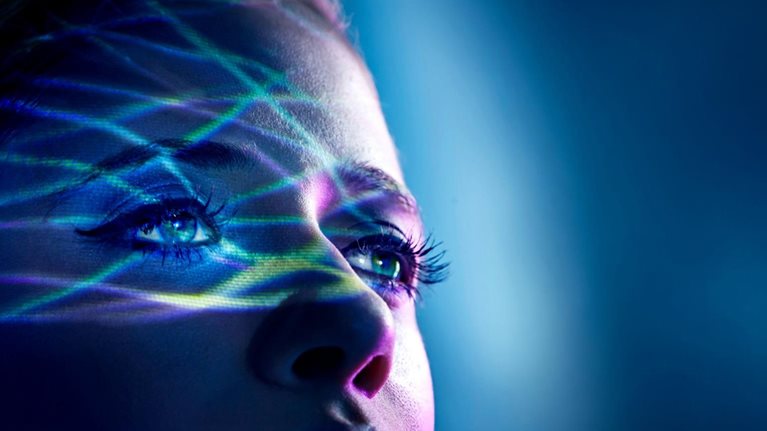 Close-up of a person's face illuminated by colorful digital light patterns, looking upward.