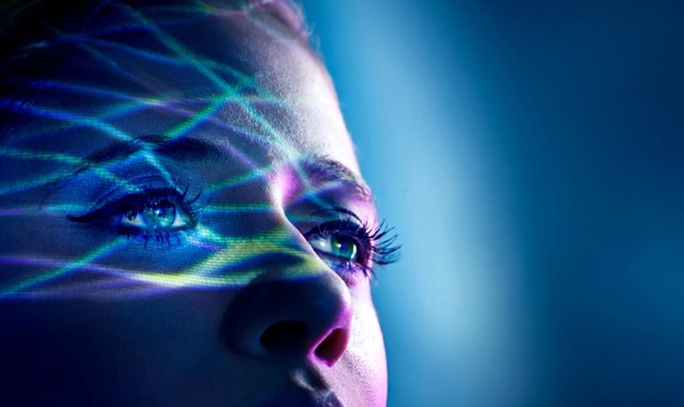 Close-up of a person's face illuminated by colorful digital light patterns, looking upward.