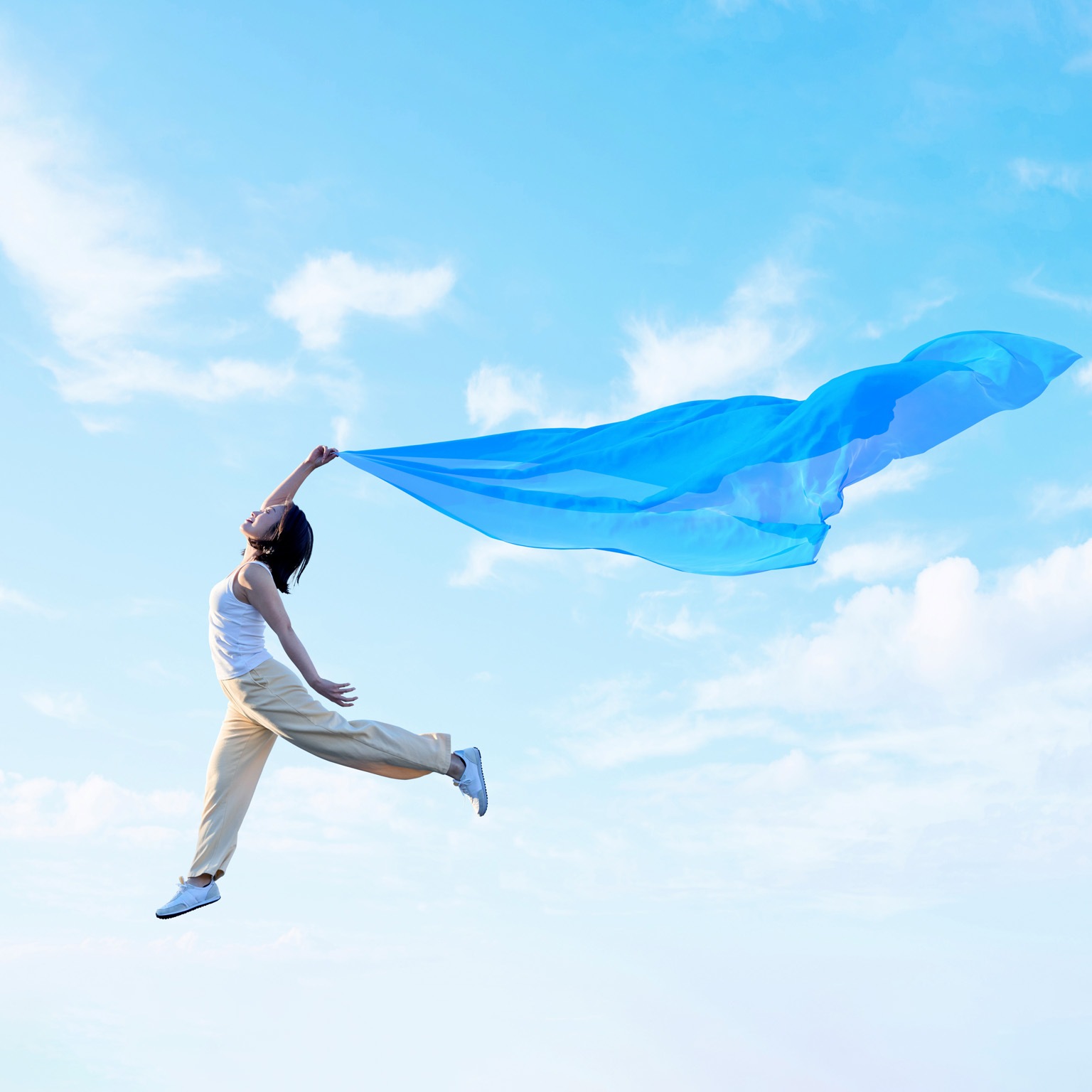 A young woman joyfully leaps through the air, holding a flowing blue scarf that billows dramatically against a bright blue sky with soft clouds.