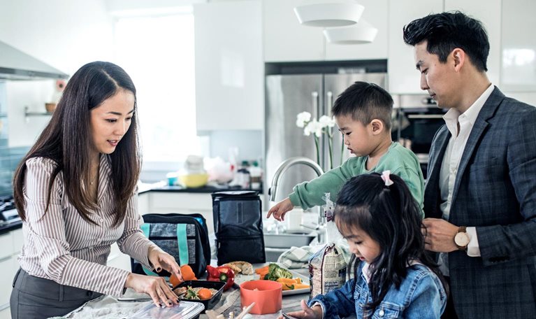 A family gathered in a bright modern kitchen, with parents and two children preparing food and packing containers together.